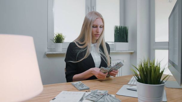 Female counting money at office workplace. Beautiful young blond woman in business suit sitting in office at light wooden desk with computer concentrating on counting large pile of cash in hands - Valokuva, kuva