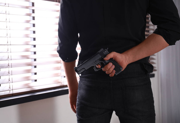 Man with gun near window indoors, closeup - Фото, изображение