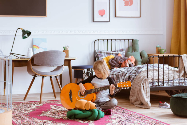 Cute blond girl sitting on the floor of her trendy bedroom,learning how to play a guitar - Foto, Imagen