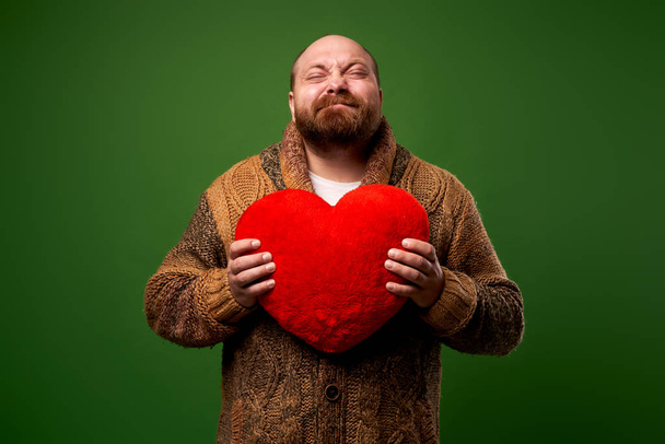 Man with closed eyes holds toy heart while standing on empty green background - Foto, imagen