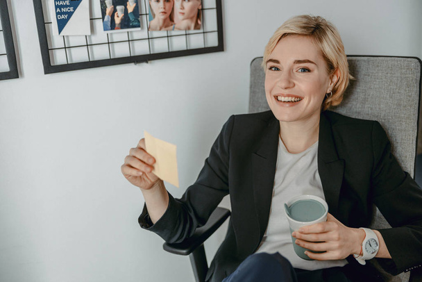 Smiling pretty woman holding hot drink while resting at workplace - Foto, Bild