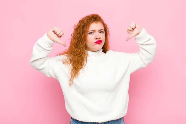 young red head woman looking sad, disappointed or angry, showing thumbs down in disagreement, feeling frustrated against pink wall - Photo, image
