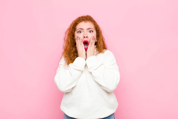 young red head woman feeling shocked and scared, looking terrified with open mouth and hands on cheeks against pink wall - Фото, изображение