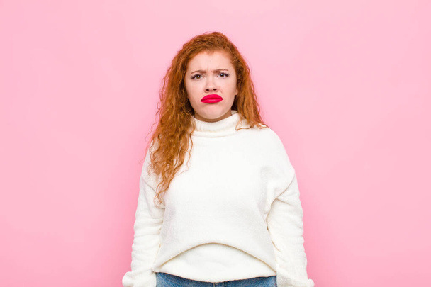 young red head woman feeling sad and whiney with an unhappy look, crying with a negative and frustrated attitude against pink wall - Foto, afbeelding