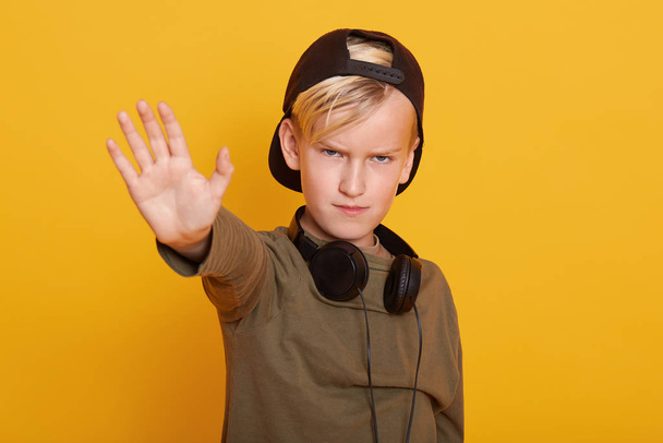 Portrait of little cute boy with angry facial expression, wearing sweater and cap, having headphones around neck, showing stop gesture with his palm, isolated over yelow wall. People emotions concept. - Photo, image