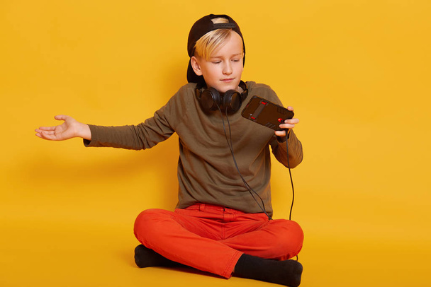 Image of little boy wearing in green sweater and blackcap, holding mobile phone in one hand, spreading other hand aside, squeezing shoulders, plays game on smart phone isolated over yellow background. - Foto, Imagen