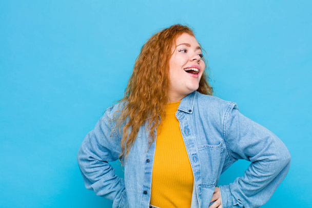 young red head woman looking happy, cheerful and confident, smiling proudly and looking to side with both hands on hips against blue wall - Фото, зображення