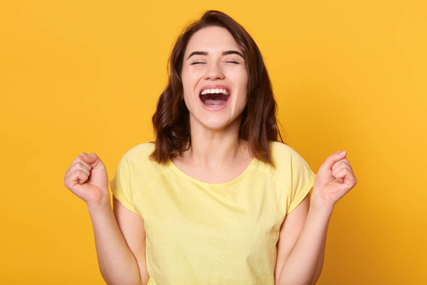 Close up portraiot of happy young woman with beautiful dark hair, clenching fist while looking directly at camera, wearing casual t shirt, isolated over yellow background. People emotions concept. - Foto, Bild