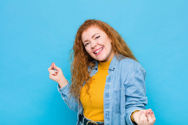 young red head woman smiling, feeling carefree, relaxed and happy, dancing and listening to music, having fun at a party against blue wall - Photo, image