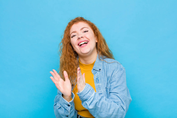 young red head woman feeling happy and successful, smiling and clapping hands, saying congratulations with an applause against blue wall - Foto, imagen