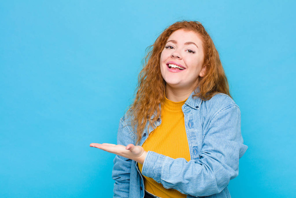 young red head woman smiling cheerfully, feeling happy and showing a concept in copy space with palm of hand against blue wall - Фото, зображення