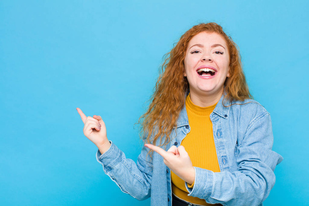 young red head woman smiling happily and pointing to side and upwards with both hands showing object in copy space against blue wall - Foto, imagen