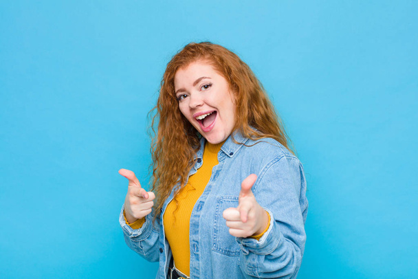 young red head woman feeling happy, cool, satisfied, relaxed and successful, pointing at camera, choosing you against blue wall - 写真・画像