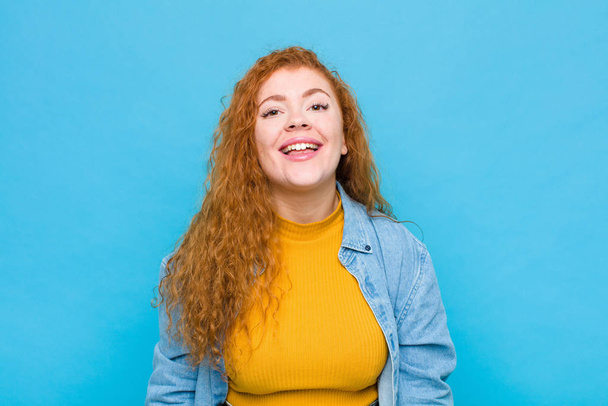 young red head woman with a big, friendly, carefree smile, looking positive, relaxed and happy, chilling against blue wall - Fotografie, Obrázek