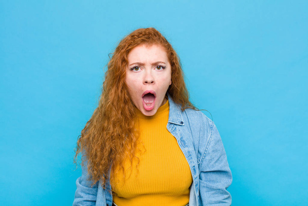 young red head woman looking shocked, angry, annoyed or disappointed, open mouthed and furious against blue wall - Foto, afbeelding