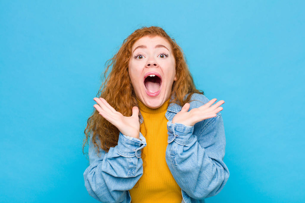 young red head woman feeling shocked and excited, laughing, amazed and happy because of an unexpected surprise against blue wall - Photo, Image