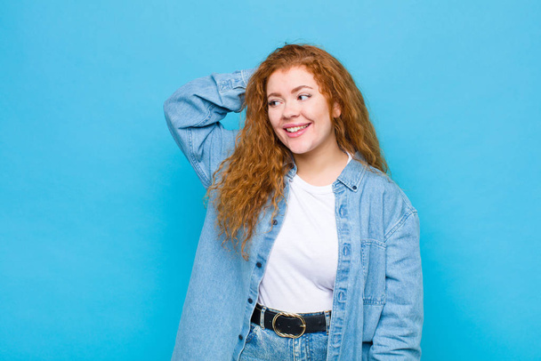 young red head woman smiling cheerfully and casually, taking hand to head with a positive, happy and confident look against blue wall - 写真・画像