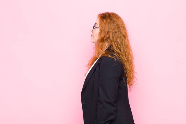young red head woman on profile view looking to copy space ahead, thinking, imagining or daydreaming against pink wall - Photo, Image