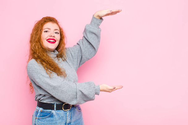 young red head woman smiling, feeling happy, positive and satisfied, holding or showing object or concept on copy space against pink wall - Photo, image