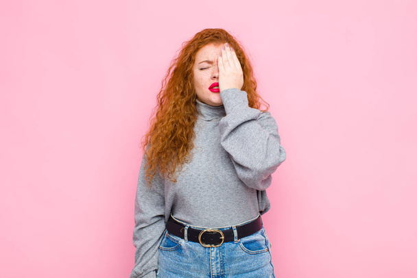 young red head woman looking sleepy, bored and yawning, with a headache and one hand covering half the face against pink wall - Фото, изображение