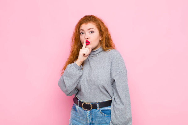 young red head woman asking for silence and quiet, gesturing with finger in front of mouth, saying shh or keeping a secret against pink wall - Fotoğraf, Görsel