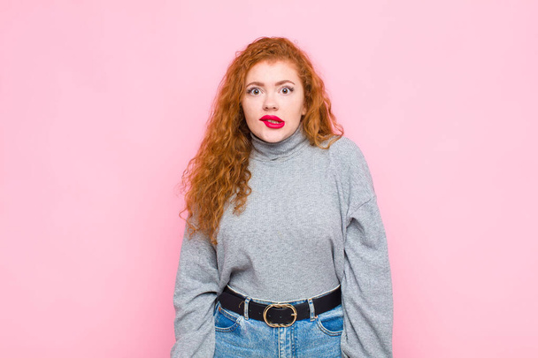 young red head woman looking puzzled and confused, biting lip with a nervous gesture, not knowing the answer to the problem against pink wall - Photo, image