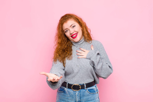 young red head woman feeling happy and in love, smiling with one hand next to heart and the other stretched up front against pink wall - 写真・画像
