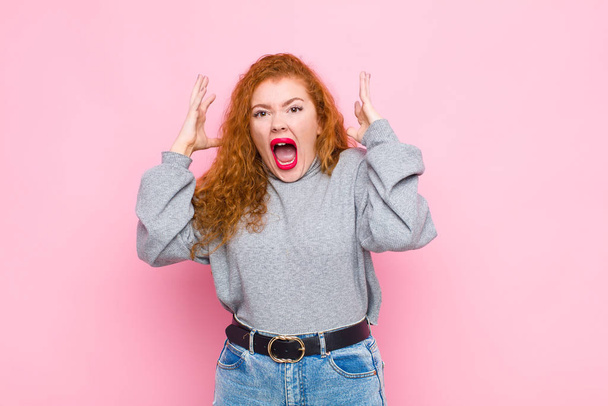 young red head woman screaming with hands up in the air, feeling furious, frustrated, stressed and upset against pink wall - Фото, зображення