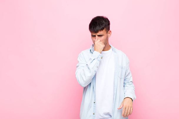 young handsome man feeling disgusted, holding nose to avoid smelling a foul and unpleasant stench against pink wall - Photo, image