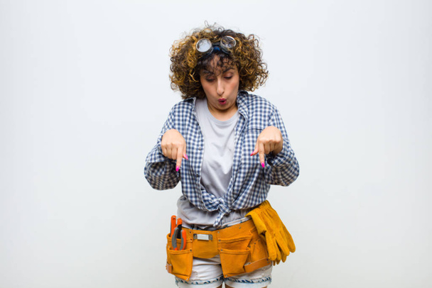 young housekeeper woman with open mouth pointing downwards with both hands, looking shocked, amazed and surprised against white wall - Foto, Imagen