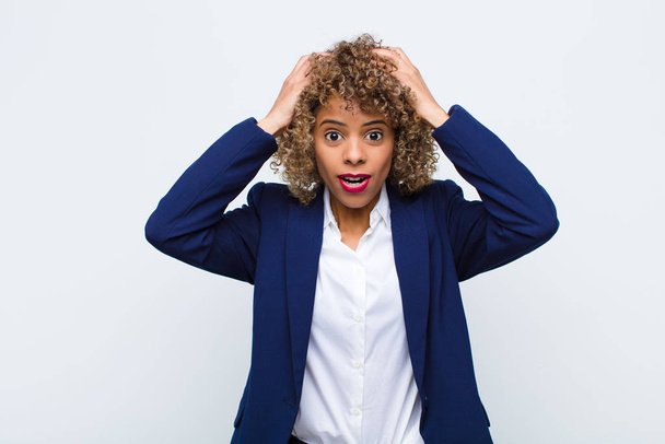 young woman african american looking excited and surprised, open-mouthed with both hands on head, feeling like a lucky winner against flat wall - 写真・画像