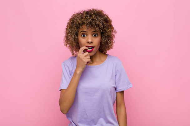 young african american woman with surprised, nervous, worried or frightened look, looking to the side towards copy space against pink wall - Photo, Image