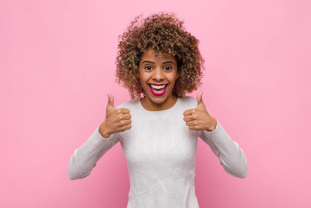 young african american woman smiling broadly looking happy, positive, confident and successful, with both thumbs up against pink wall - Foto, imagen