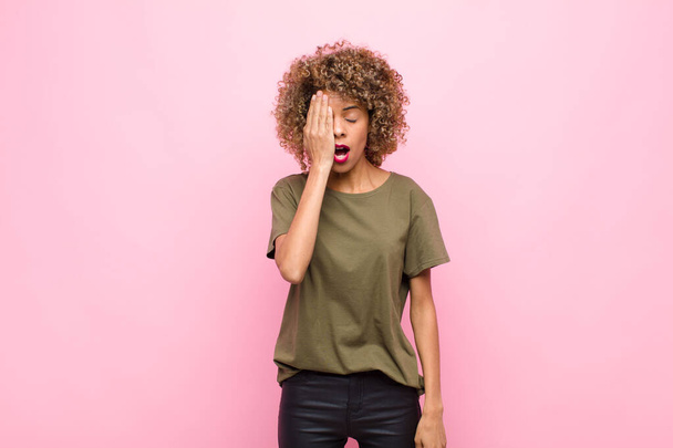young african american woman looking sleepy, bored and yawning, with a headache and one hand covering half the face against pink wall - Foto, imagen