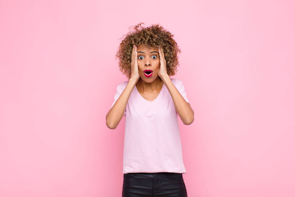jeune femme afro-américaine regardant désagréablement choqué, effrayé ou inquiet, bouche grande ouverte et couvrant les deux oreilles avec les mains contre le mur rose
 - Photo, image