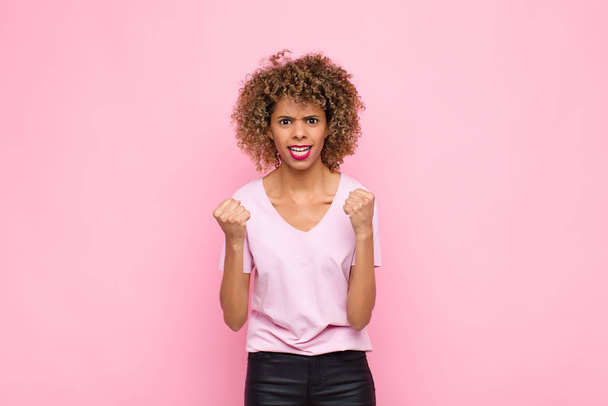 young african american woman shouting triumphantly, laughing and feeling happy and excited while celebrating success against pink wall - Photo, image