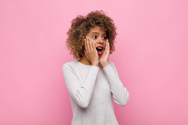 young african american woman feeling happy, excited and surprised, looking to the side with both hands on face against pink wall - Photo, Image