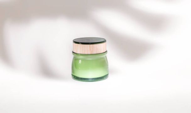 Cream in a green jar on a white background under the shadow of a tropical monstera plant. Eco cosmetics concept - Foto, Imagen