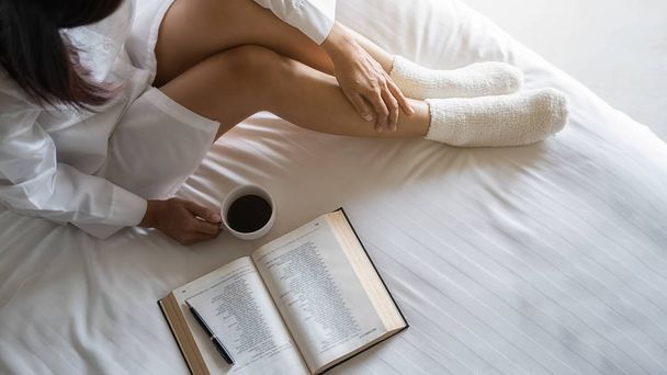 Азиатка, держащая чашку кофе и читающая книгу на кровати после утреннего пробуждения. Утренний стиль жизни. - Фото, изображение