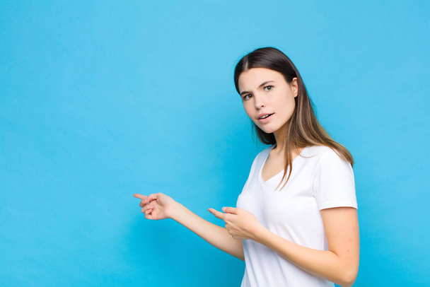 young pretty woman feeling shocked and surprised, pointing to copy space on the side with amazed, open-mouthed look against blue wall - 写真・画像