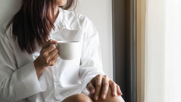 Азиатка, держащая чашку кофе и сидящая у окна после утреннего пробуждения. Утренний стиль жизни
. - Фото, изображение