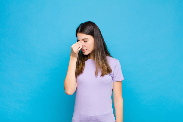 young pretty woman feeling stressed, unhappy and frustrated, touching forehead and suffering migraine of severe headache against blue wall - Photo, Image