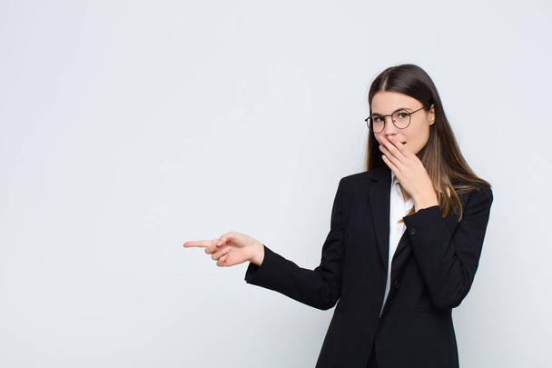 young businesswoman feeling happy, shocked and surprised, covering mouth with hand and pointing to lateral copy space against white wall - Foto, Imagem