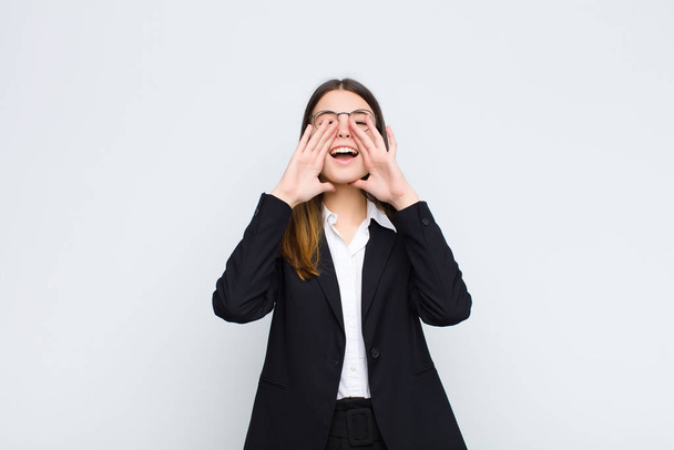 young businesswoman feeling happy, excited and positive, giving a big shout out with hands next to mouth, calling out against white wall - Foto, Imagem