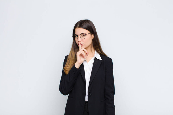 young businesswoman asking for silence and quiet, gesturing with finger in front of mouth, saying shh or keeping a secret against white wall - Foto, afbeelding