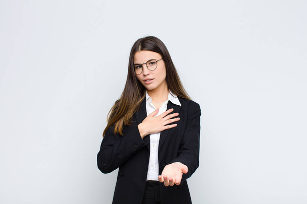 young businesswoman feeling happy and in love, smiling with one hand next to heart and the other stretched up front against white wall - Fotografie, Obrázek