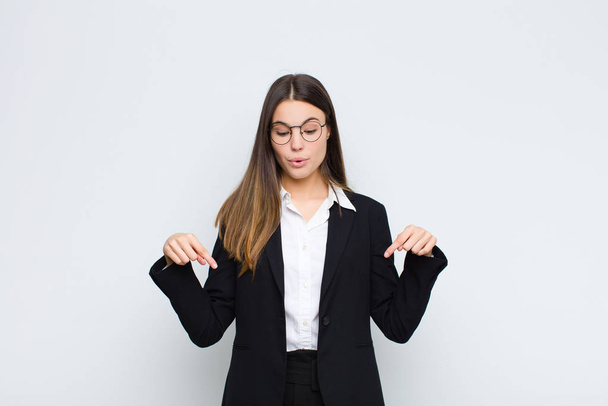 young businesswoman feeling shocked, open-mouthed and amazed, looking and pointing downwards in disbelief and surprise against white wall - Photo, image