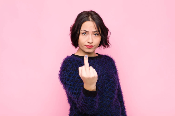young pretty woman feeling angry, annoyed, rebellious and aggressive, flipping the middle finger, fighting back against pink wall - Foto, Imagen