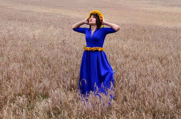 Una joven está en un campo de trigo con un vestido azul. Tiene una corona de flores silvestres amarillas en su cabeza. En la cintura tiene un cinturón de flores amarillas
. - Foto, Imagen