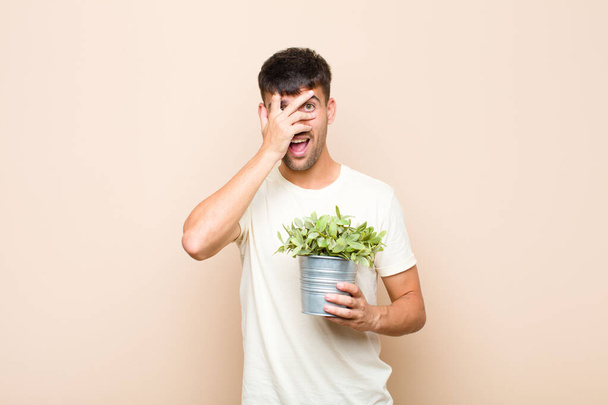 young handsome man looking shocked, scared or terrified, covering face with hand and peeking between fingers holding a plant - Foto, Bild